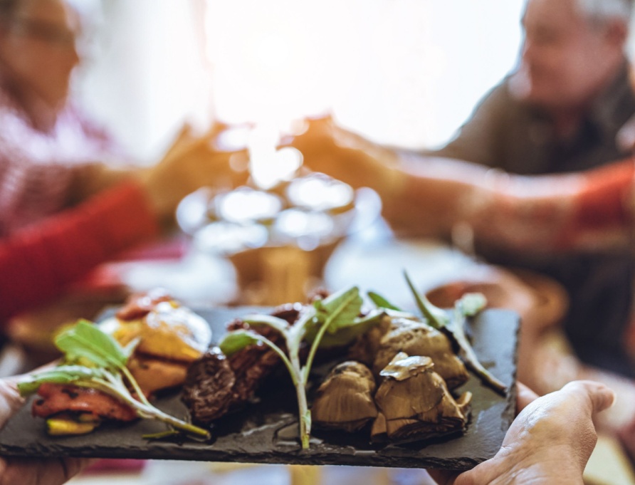 Chef bringing out platter of food, with group of people at a table in the background