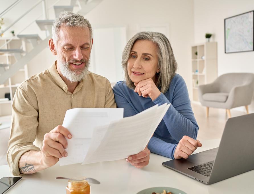 A senior couple sit at a laptop comparing multiple documents